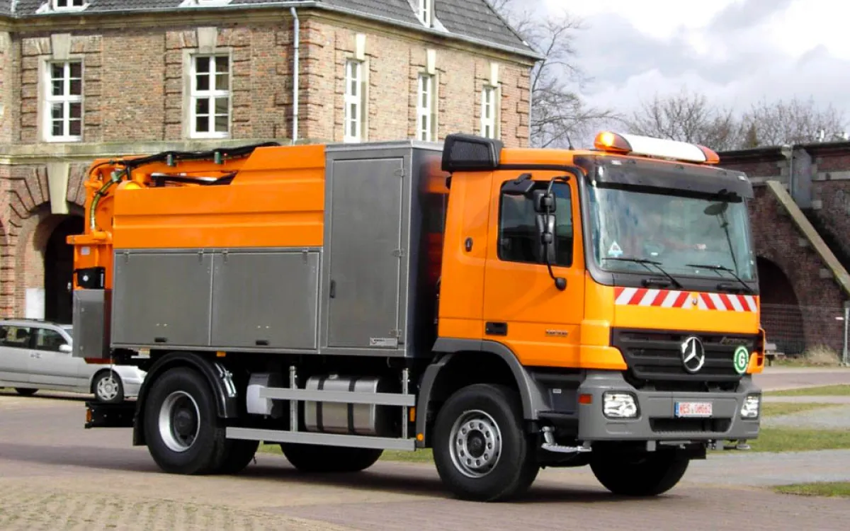 Orangefarbenes HD-Spülfahrzeug auf Mercedes-Fahrgestell mit Edelstahl-Geräteschränken vor einem historischen Backsteingebäude